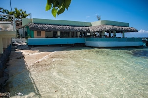 Um edifício à beira-mar com telhado de palha, piscina e águas claras e rasas.
