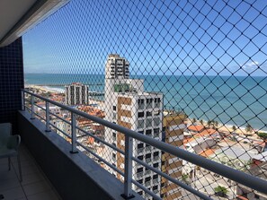 Un balcón con barandilla de malla metálica que da a una vista urbana costera, con edificios modernos y una vista despejada del océano.
