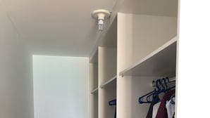 Un couloir étroit avec une valise, une armoire où sont suspendus des vêtements et un luminaire au plafond.