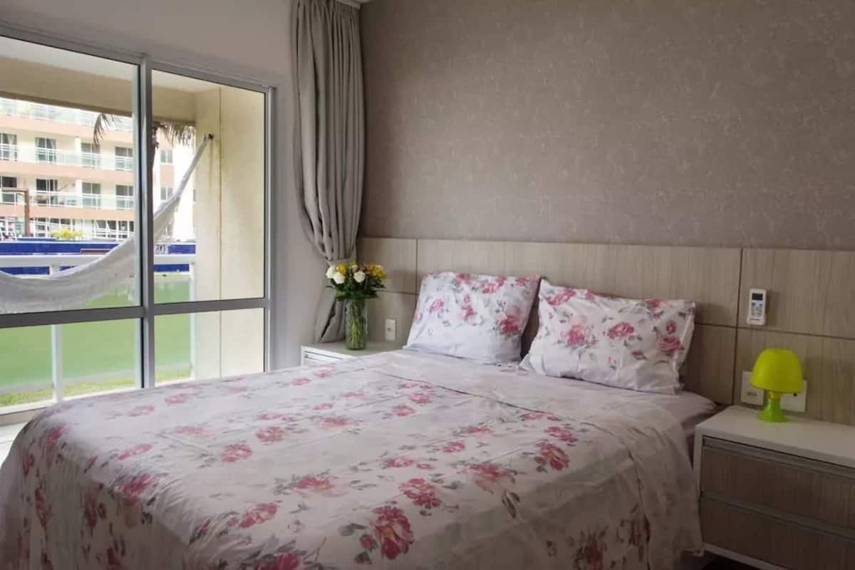 Quarto de hotel com uma cama grande, uma mesa de cabeceira com abajur, um vaso com flores e vista para um edifício pela janela.