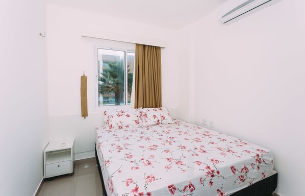 A bedroom with a bed, a nightstand, a window with curtains, and a view of palm trees outside. - VG FUN Meu Lugar Ceará (Fortaleza)