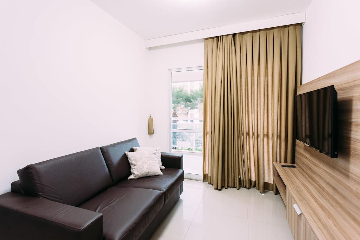 Uma sala de estar moderna com um sofá de couro marrom escuro, um rack de madeira para TV e uma janela grande com cortinas bege.