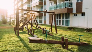 Un terrain de jeux avec des balançoires et un toboggan, situé devant un immeuble d’appartements moderne.