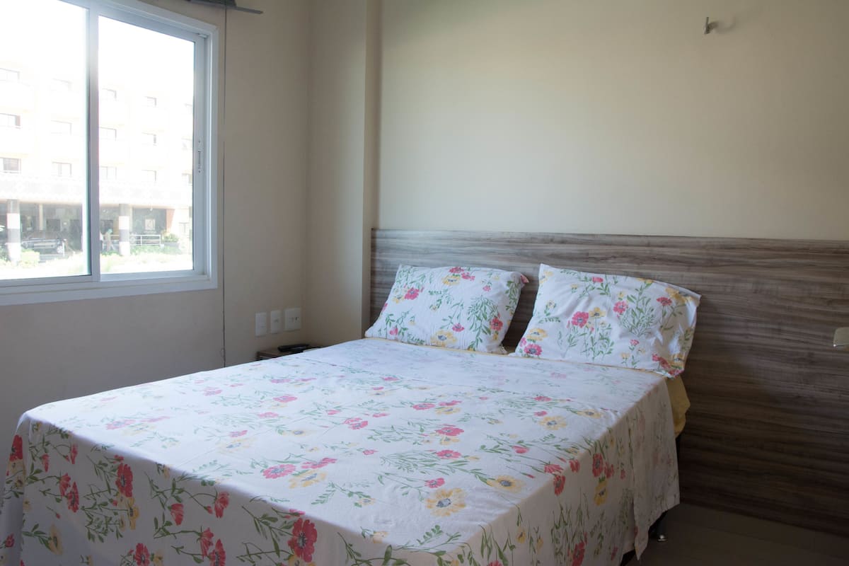 Quarto com cama, colcha florida e janela com vista para edifícios.