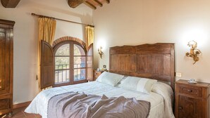 A bedroom with a wooden headboard, a large bed with a white and brown quilt, a nightstand with a lamp, and a view of the outdoors through a window with curtains. - Tenuta La Pieve Casale La Valle (Pergine Valdarno)