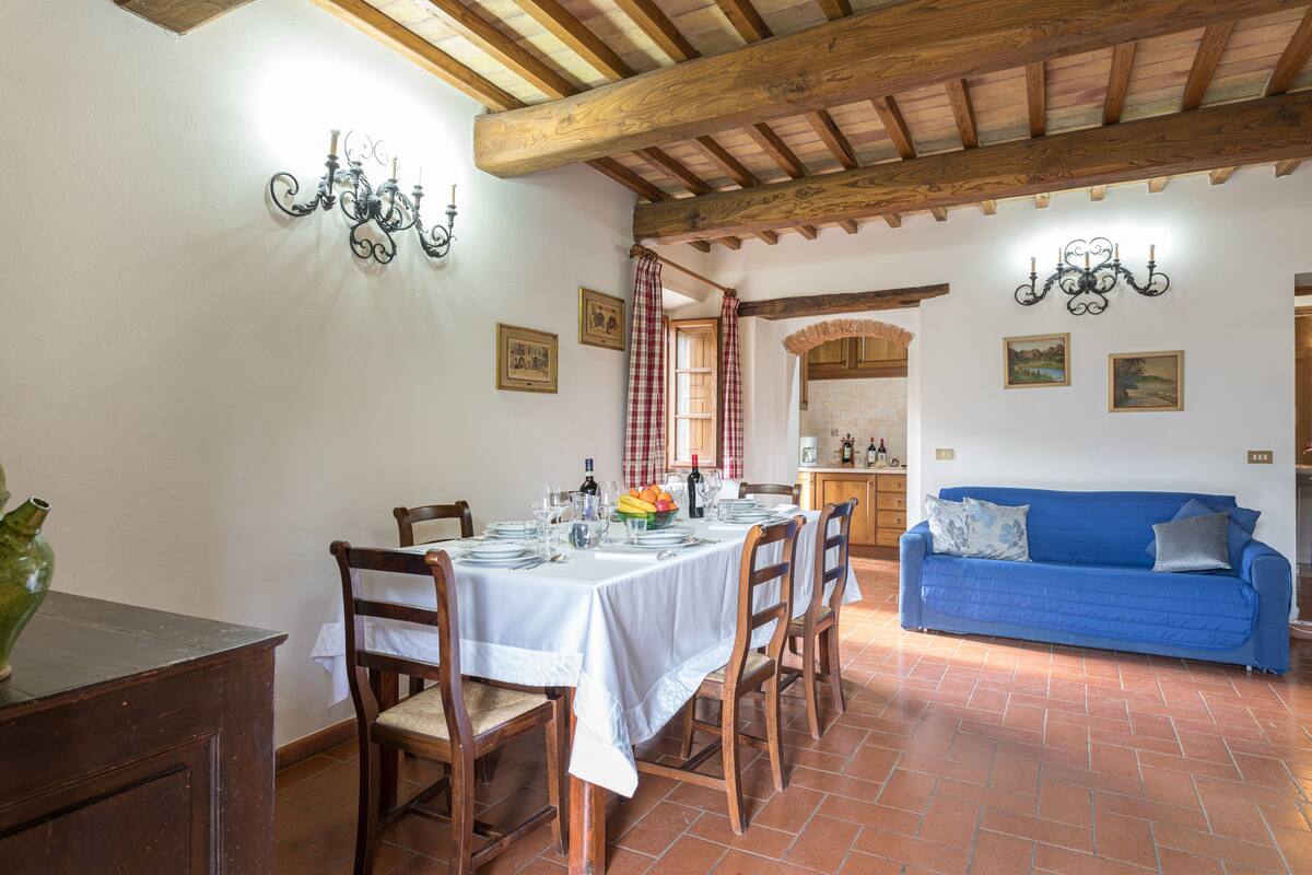 Un coin repas avec une table dressée pour quatre personnes, des chaises en bois, un canapé bleu et, au fond, une cuisine.