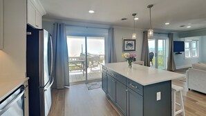 A modern kitchen with a central island, a refrigerator, and a dining area with a view. - Kirk's Mema's Beach | Pet Friendly + Walk to Beach (Topsail Beach)