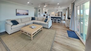 A modern living room with a sofa, coffee table, and a kitchen area in the background. - Kirk's Mema's Beach | Pet Friendly + Walk to Beach (Topsail Beach)