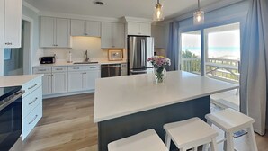A modern kitchen with white cabinets, a large island with bar stools, a refrigerator, and a view of the ocean through large windows. - Kirk's Mema's Beach | Pet Friendly + Walk to Beach (Topsail Beach)