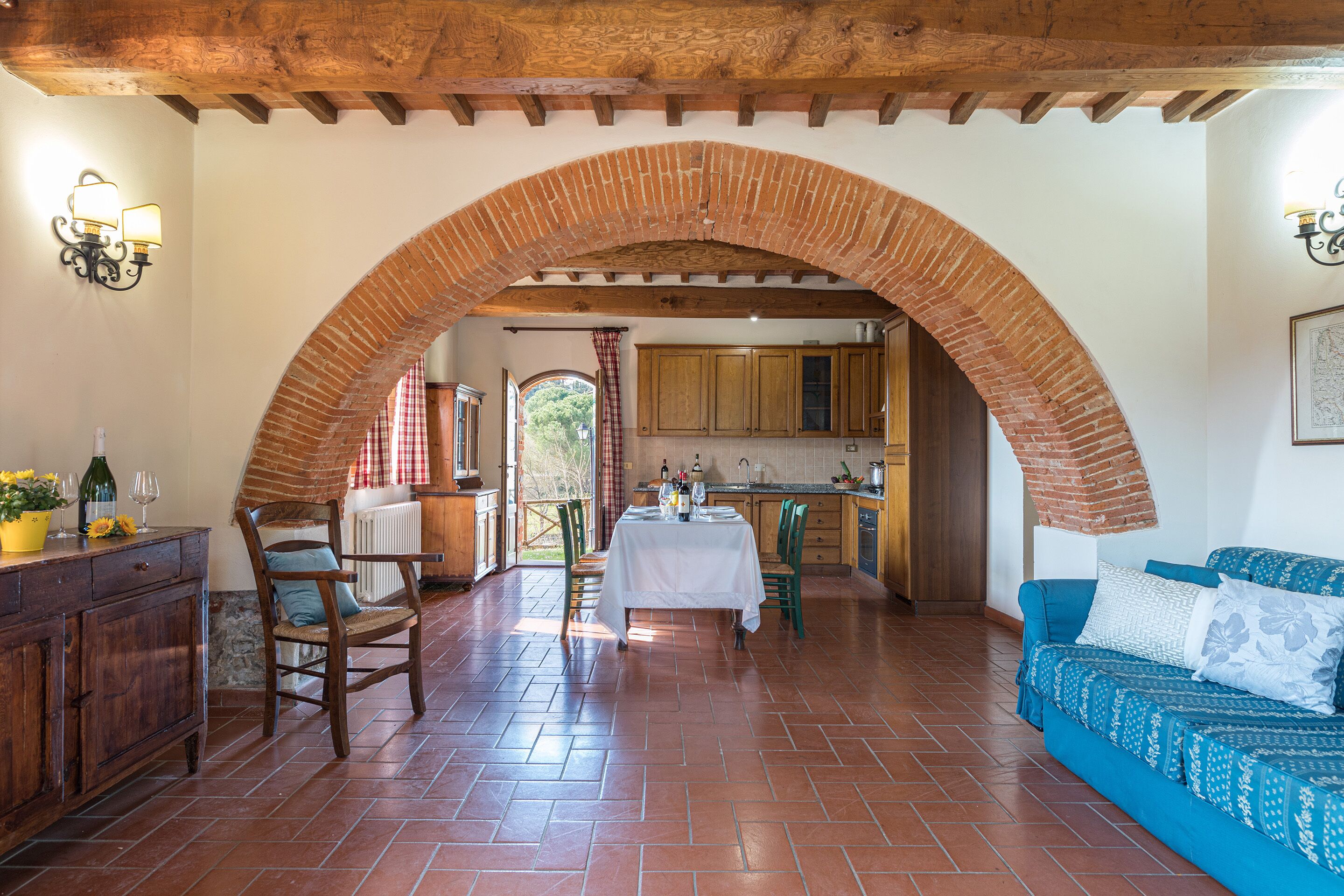 A spacious dining area with a brick archway, wooden ceiling beams, and a view into the kitchen.