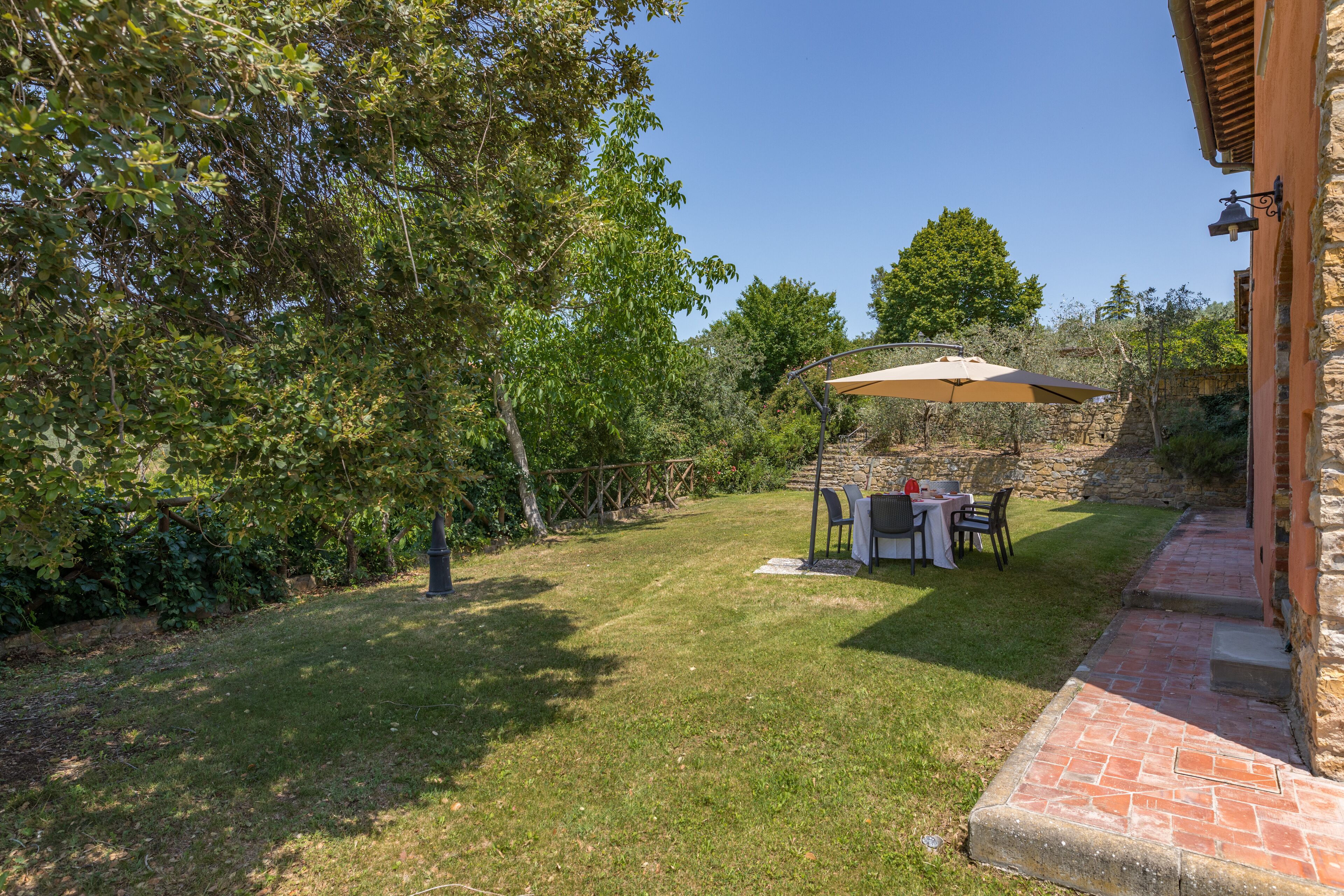 A patio area with a table and chairs under a canopy, surrounded by a stone wall and greenery.