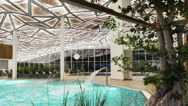 An indoor swimming pool with a geometric ceiling structure, surrounded by indoor plants and seating areas. - Rezort Gothal Apartmány a Penzióny (Liptovská Osada)