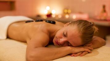 A person lying on a massage table with stones on their back, in a room with soft lighting and candles in the background.