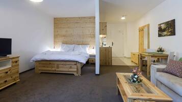 A room with a wooden bed, a television on a wooden dresser, a wooden coffee table with a flower arrangement, and a grey sofa.