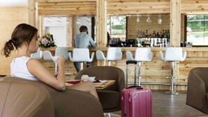 A woman sitting on a brown leather couch with a pink suitcase beside her, in a room with wooden walls and a bar area in the background. - Rezort Gothal Apartmány a Penzióny (Liptovská Osada)