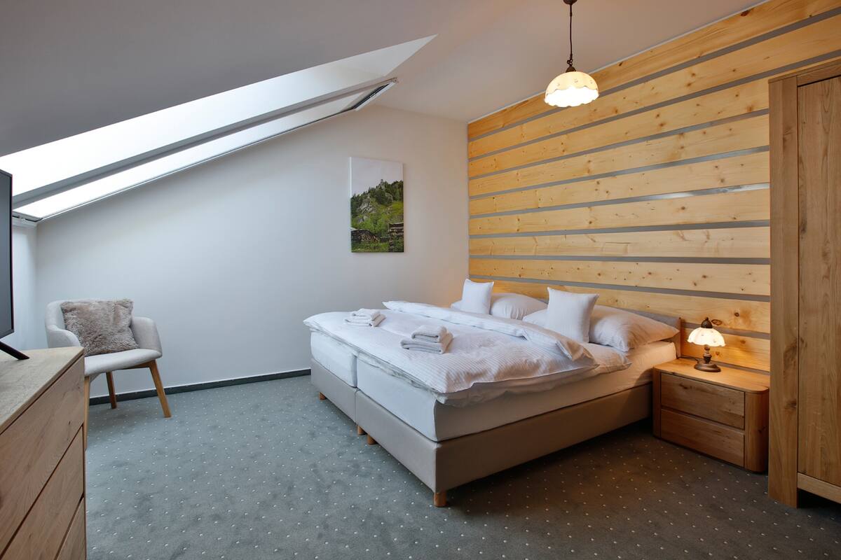 Un dormitorio con cama, una silla, una mesita de noche, una lámpara, un cuadro en la pared y un tragaluz.