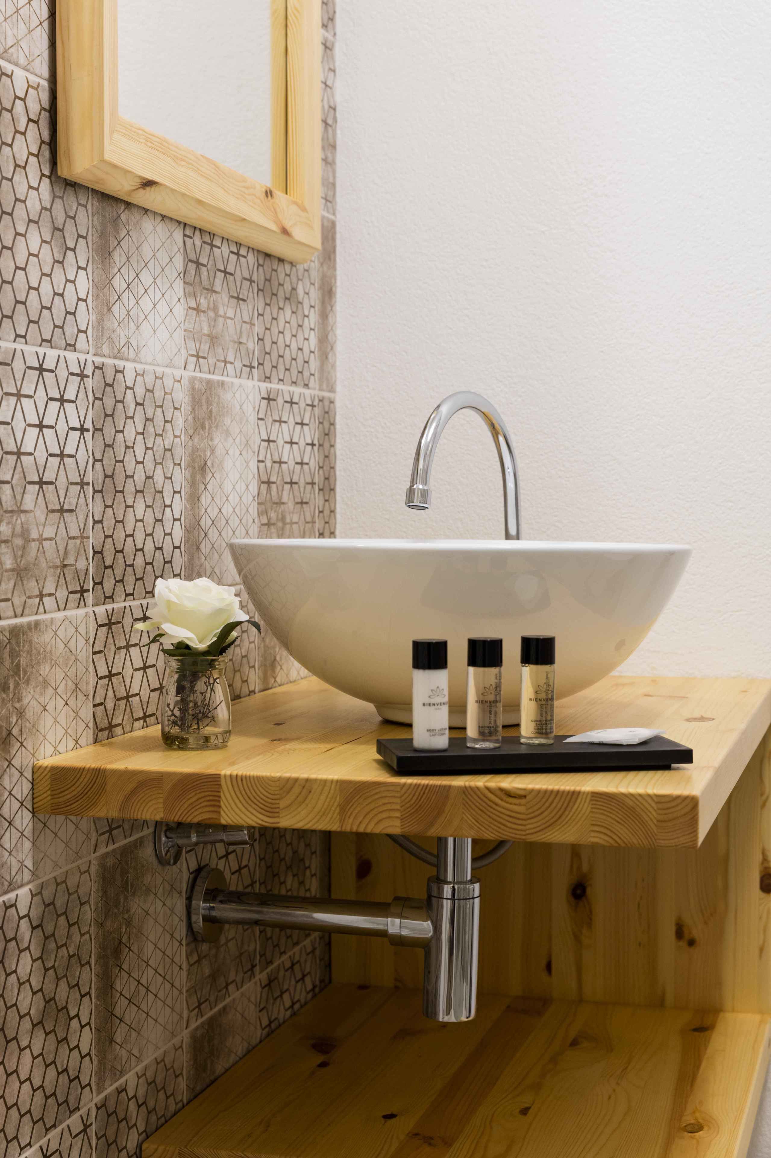 Un baño con tocador de madera, un lavabo blanco tipo cuenco y un jarrón pequeño con una flor blanca.