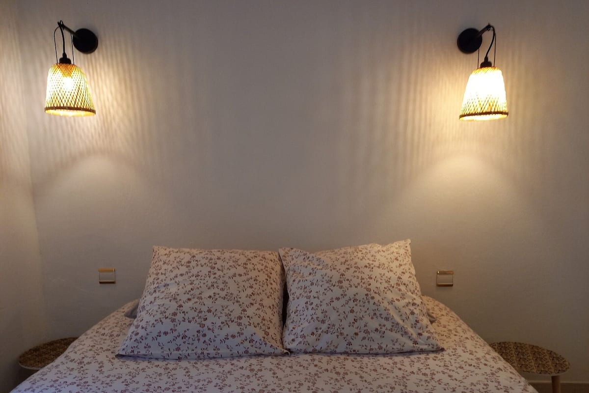 A bed with floral bedding, two wall-mounted lamps, and a small table beside the bed.