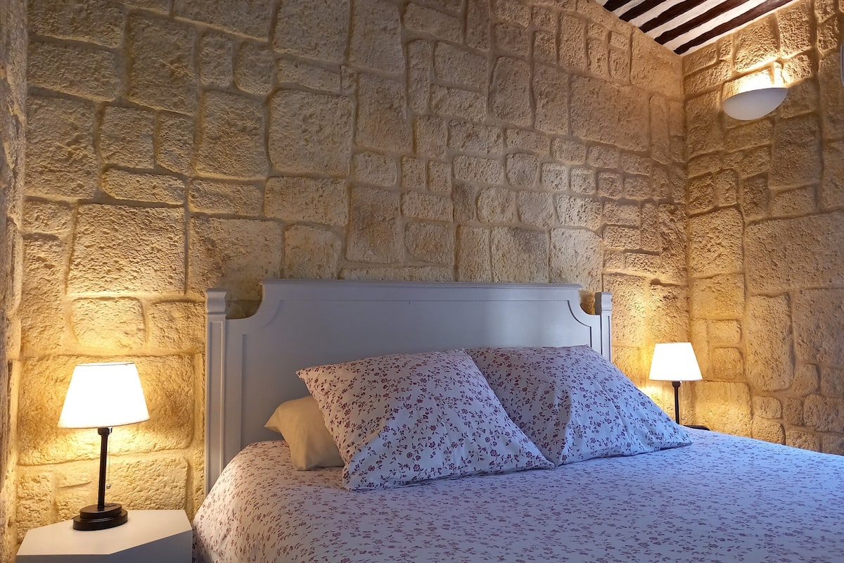 A bedroom with a stone wall, a white bed, floral bedding, and two bedside lamps.