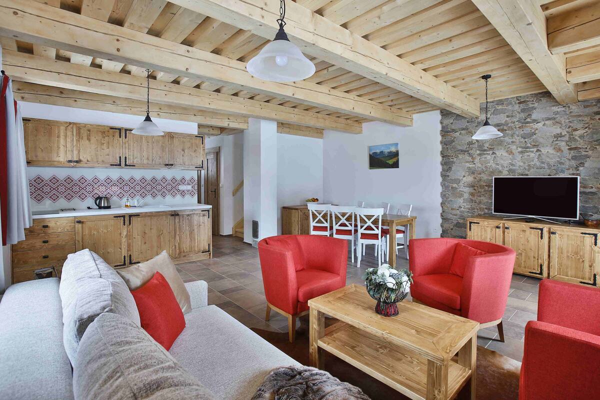 A spacious living area with a wooden ceiling, a sofa, red armchairs, a wooden coffee table, and a kitchen in the background.