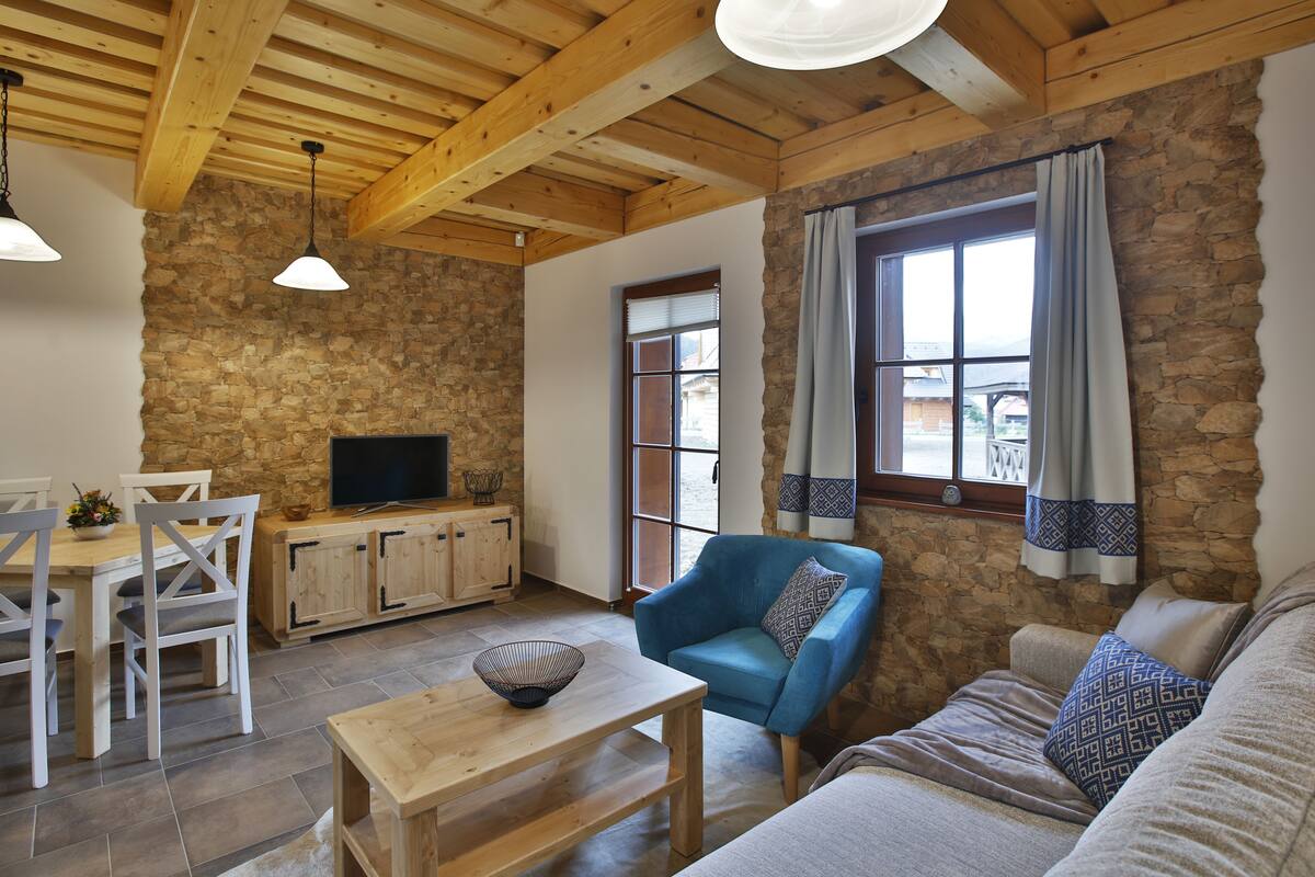 A cozy living room with a stone wall, wooden ceiling beams, a blue armchair, a sofa, a wooden coffee table, and a TV on a wooden cabinet.