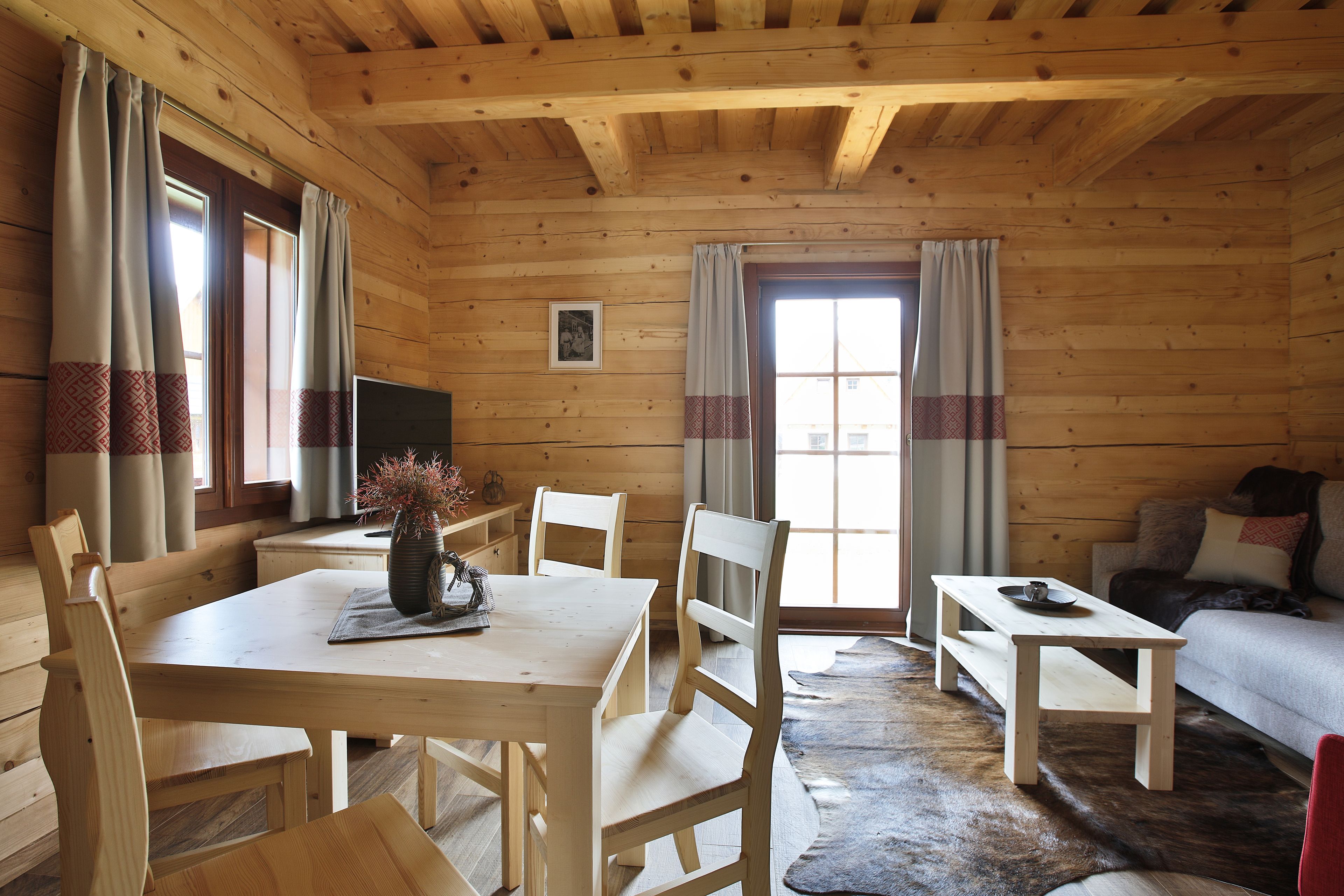 A wooden cabin interior with a dining table, chairs, a sofa, and a television.