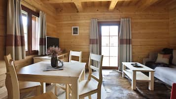 A wooden cabin interior with a dining table, chairs, a sofa, and a television.