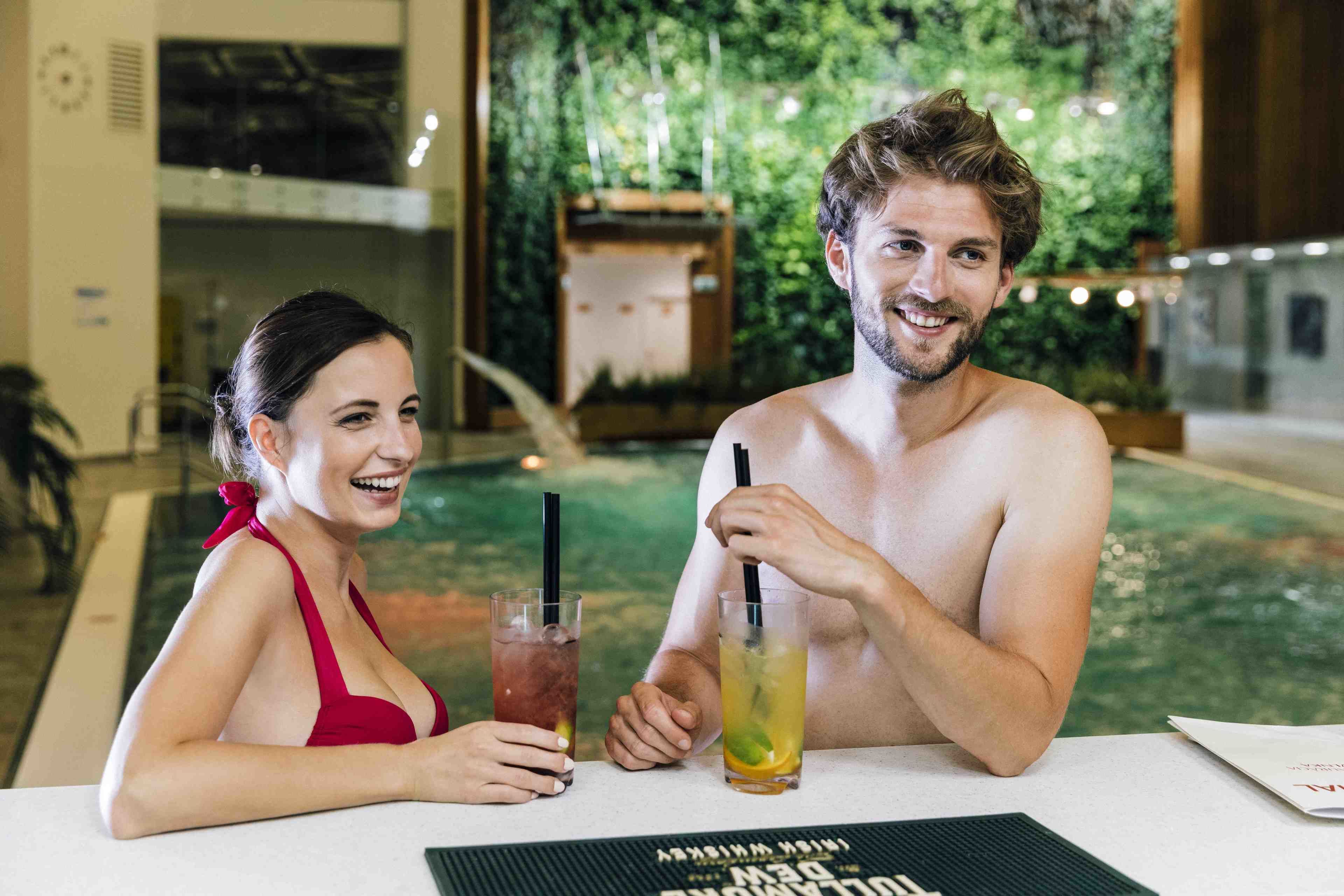 Un couple est assis à une table près d’une piscine, avec des boissons, entouré de plantes d’intérieur et d’une structure en bois.