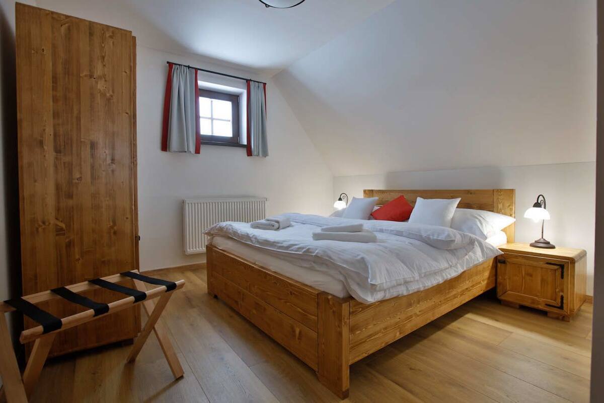 A bedroom with a wooden bed, a nightstand with a lamp, a window with curtains, and a wooden wardrobe.