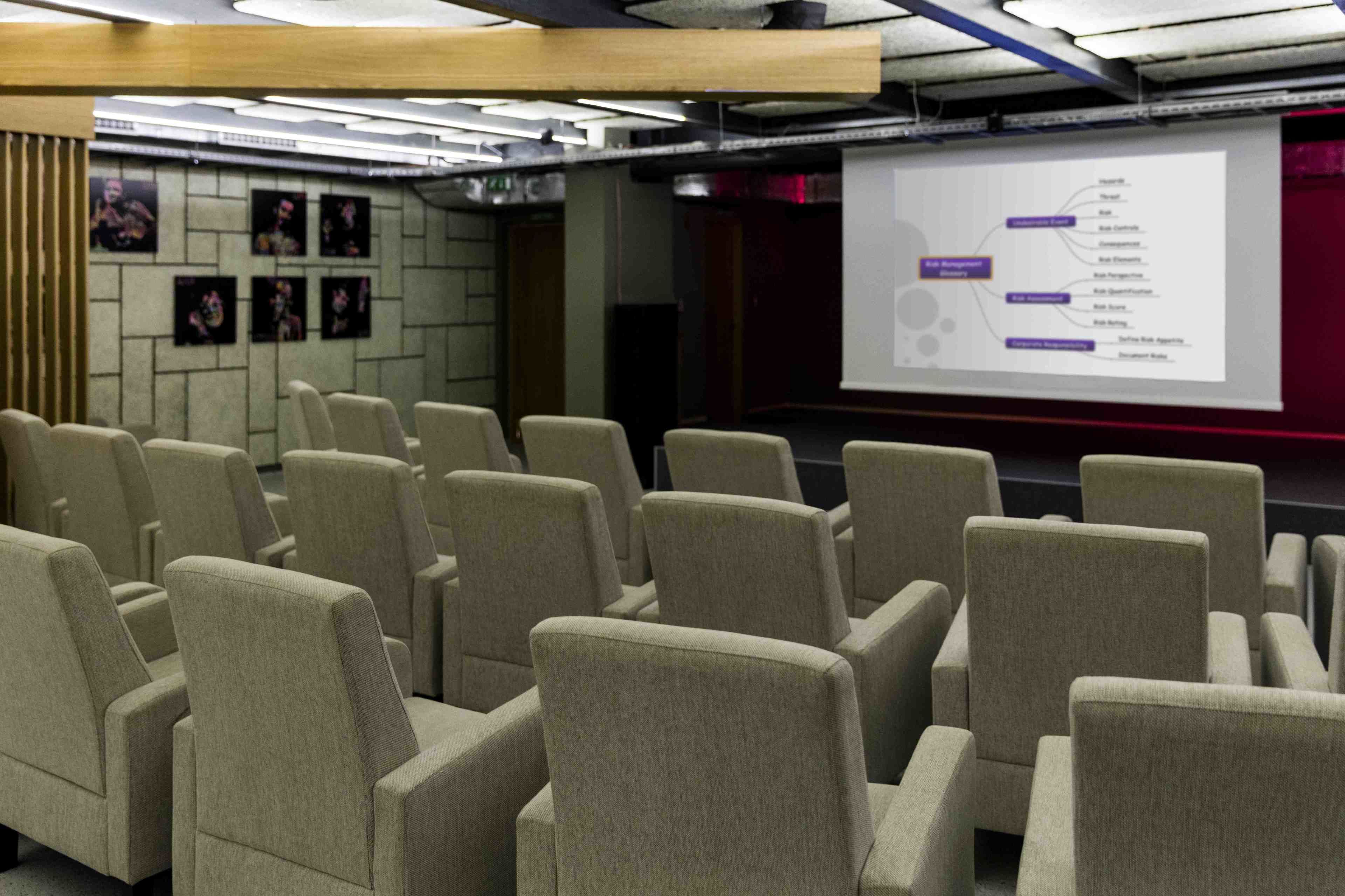 A conference room with rows of beige chairs, a projector screen displaying a presentation, and a wall with framed pictures.