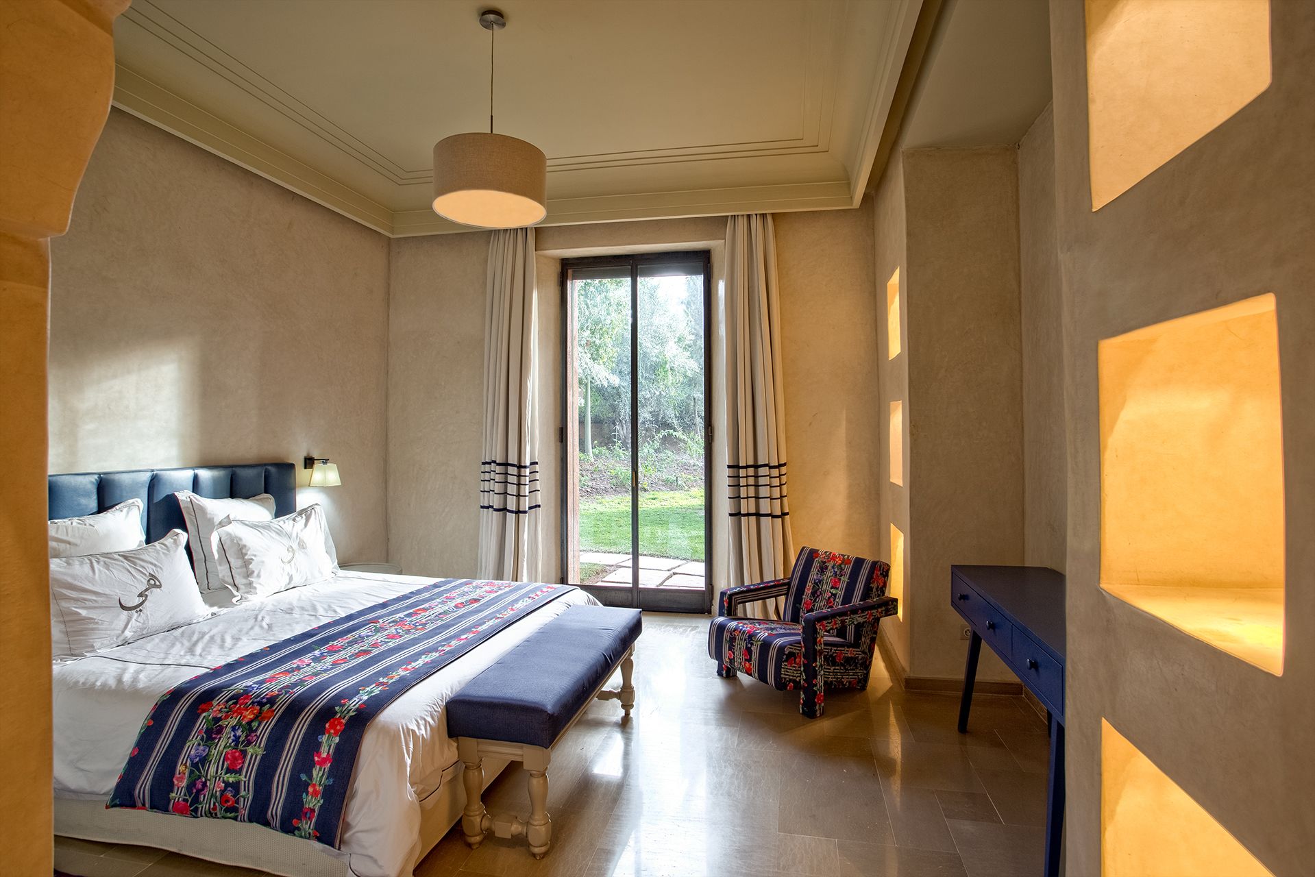 A bedroom with a large bed, a bench, a chair, and a desk. There is a window with curtains, a hanging light fixture, and a view of greenery outside.