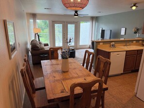 A dining area with a wooden table, chairs, a vase with flowers, a sofa, a TV, and a kitchen with a sink and cabinets.