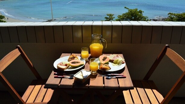 A wooden table with breakfast items and a glass of orange juice on a balcony overlooking a beach and ocean. - OP Flat Vip Frente Mar (Fortaleza)