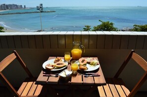 A wooden table with breakfast items and a glass of orange juice on a balcony overlooking a beach and ocean. - OP Flat Vip Frente Mar (Fortaleza)
