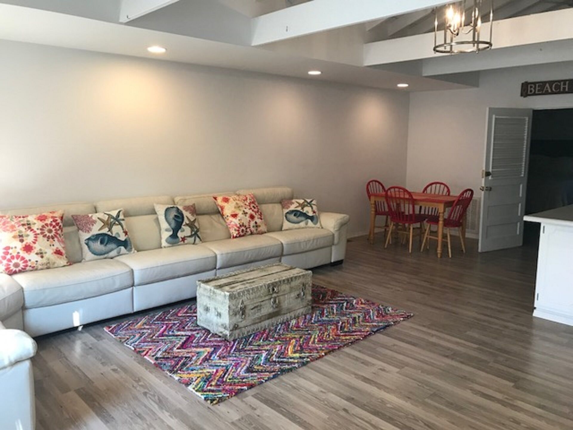 A modern living room with a sofa, colorful pillows, a patterned rug, and a dining area with red chairs.