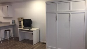 Una habitación con un escritorio blanco, televisor, armario y un taburete.