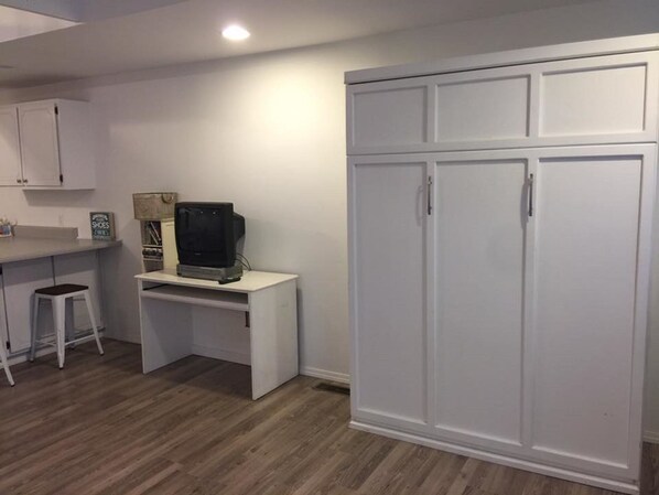Una habitación con un escritorio blanco, televisor, armario y un taburete.