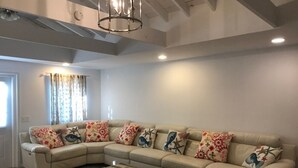 Una sala de estar con un sofá seccional color beige, cojines estampados, una mesa de centro de madera y un ventanal con cortinas.