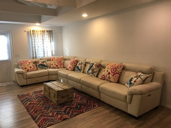 Un salon avec un canapé d’angle beige, des coussins colorés, une commode en bois et un tapis à motifs.