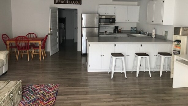Una cocina moderna con armarios blancos, una isla central con taburetes y un cartel que dice 'BEACH HOUSE'.