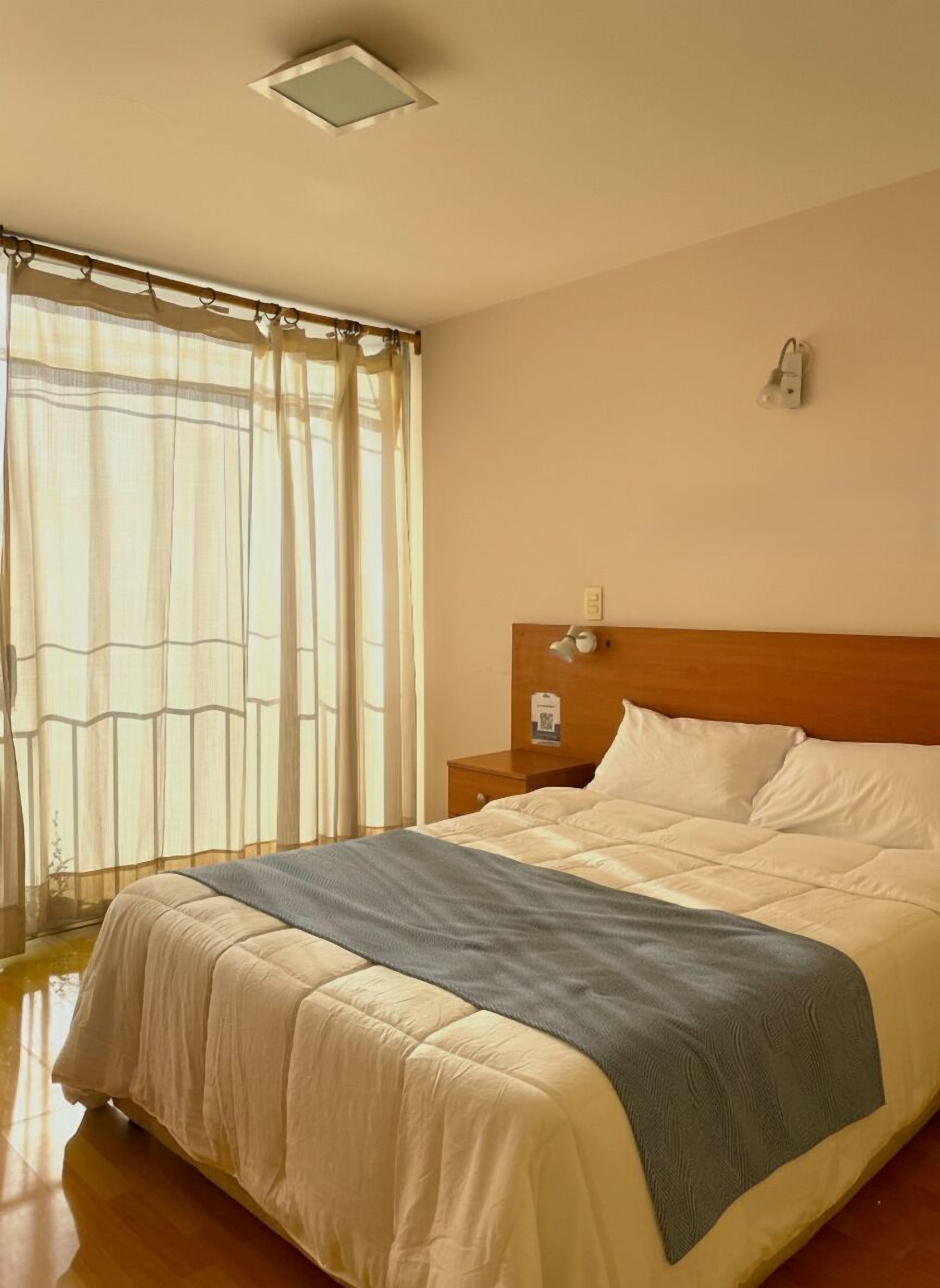 A hotel room with a bed, a nightstand, a wall-mounted lamp, and a window with curtains.