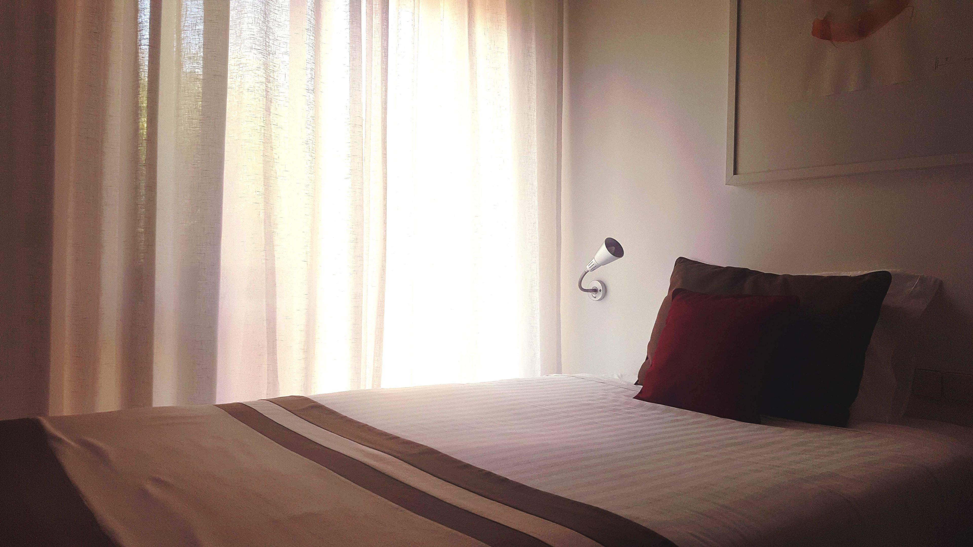 Quarto de hotel com cama, cortinas, luminária na parede e travesseiro.