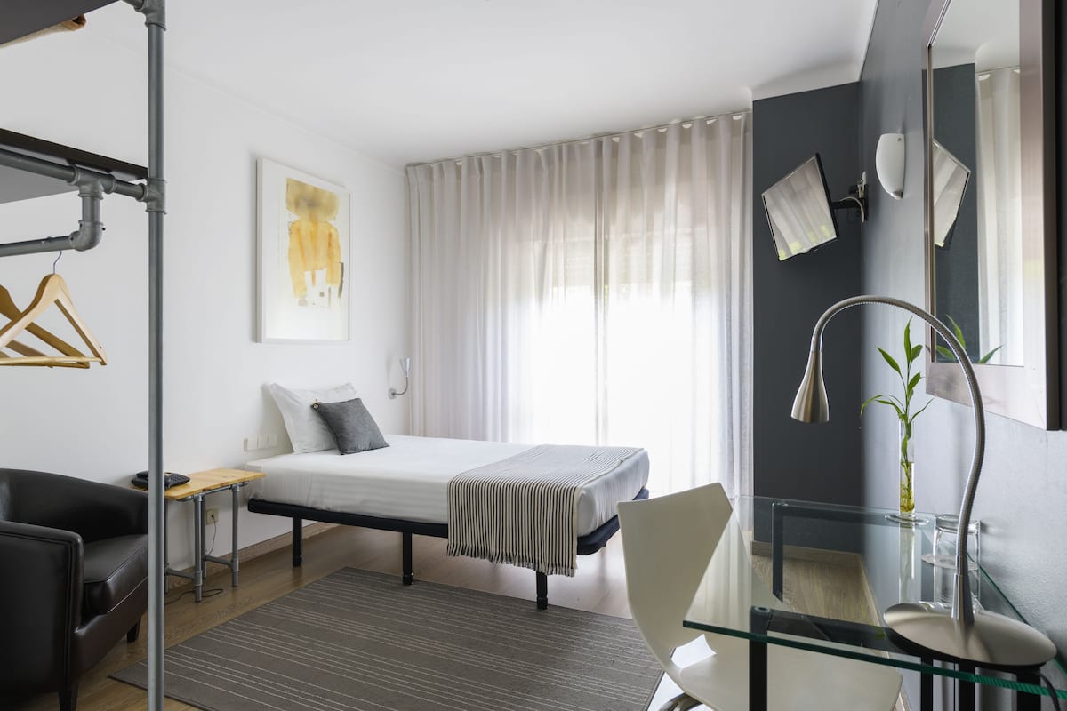 A modern bedroom with a bed, bedside table, chair, and a glass desk with a lamp.