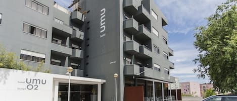Un moderno edificio de hotel con fachada gris, varios balcones y una entrada visible con un área de estar al aire libre techada.
