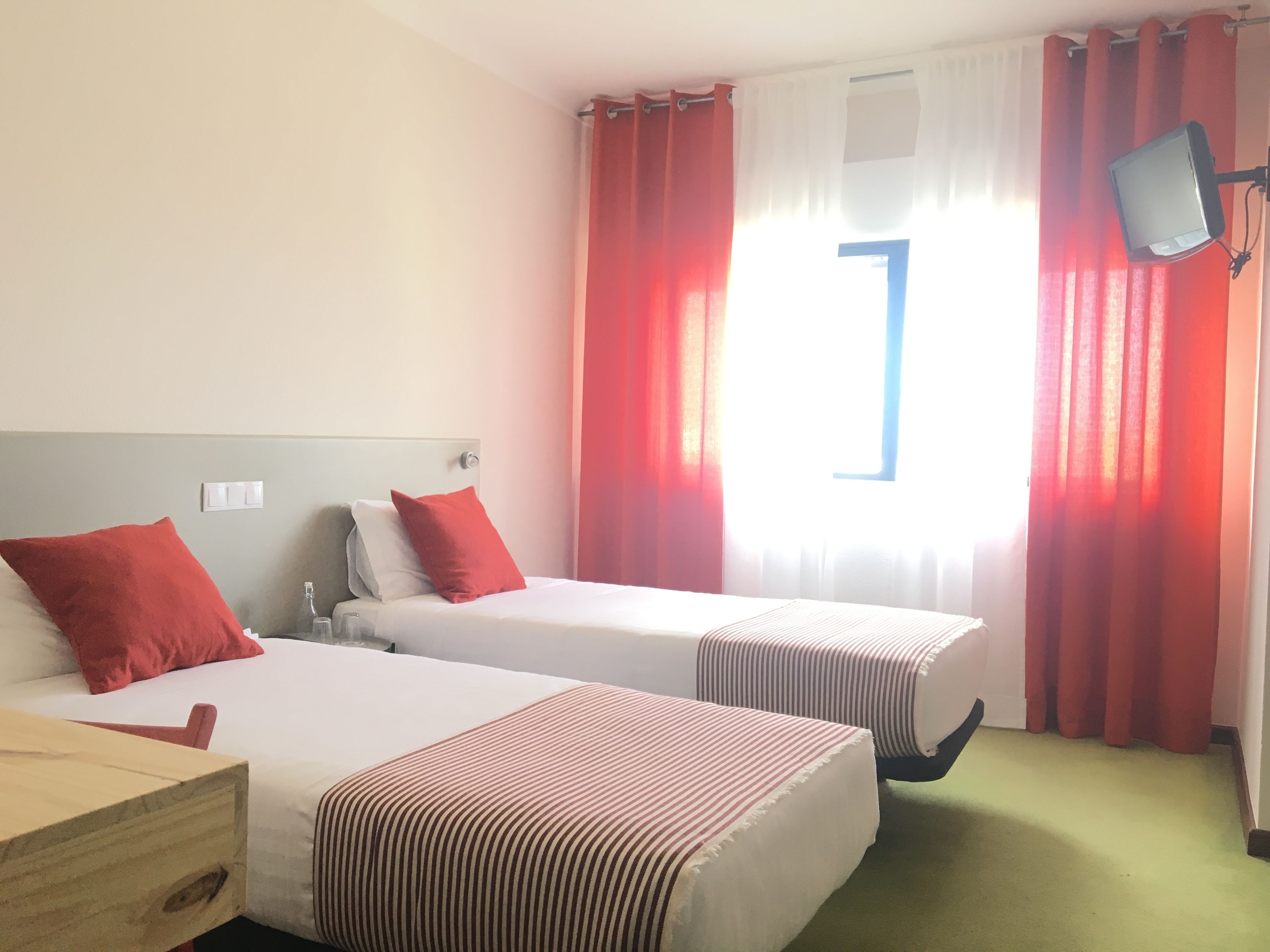 A hotel room with two beds, red and white bedding, a wooden nightstand, a TV mounted on the wall, and a window with red curtains.