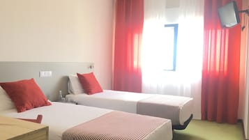 A hotel room with two beds, red and white bedding, a wooden nightstand, a TV mounted on the wall, and a window with red curtains.