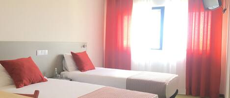 Habitación de hotel con dos camas, ropa de cama roja y blanca, una mesita de noche de madera, un televisor montado en la pared y una ventana con cortinas rojas.