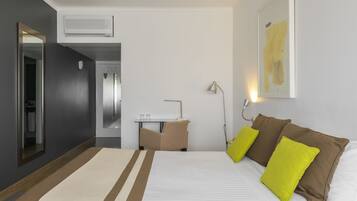 A hotel room with a bed, a desk, a chair, a mirror, a lamp, and a painting on the wall.