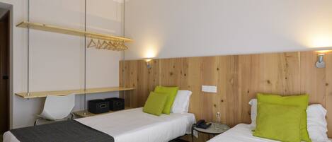 A hotel room with two beds, a wooden headboard, a small table with a phone and glass, and a wall-mounted shelf with clothes hangers.
