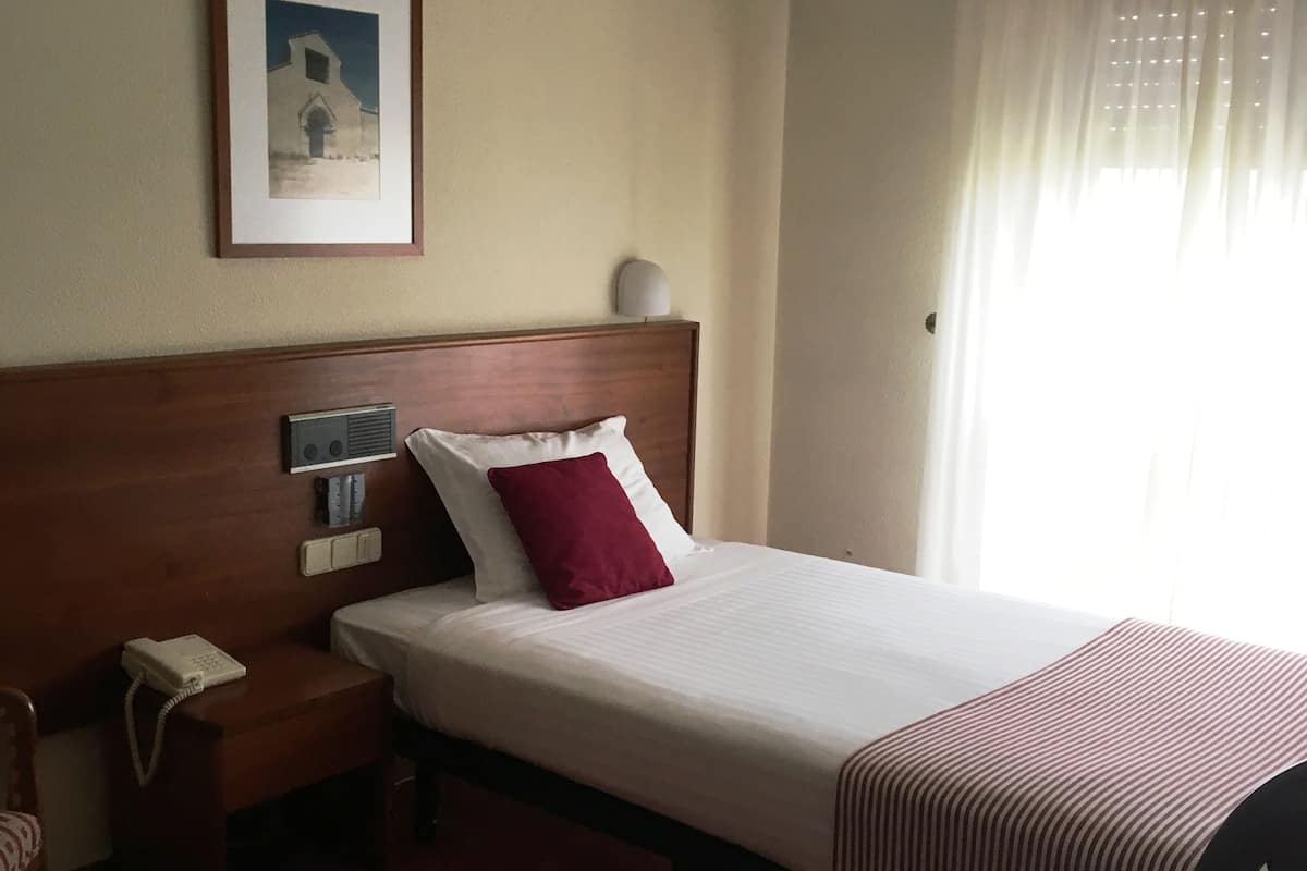 A hotel room with a bed, a bedside table with a phone, a wall-mounted light, a framed picture, and a window with sheer curtains.